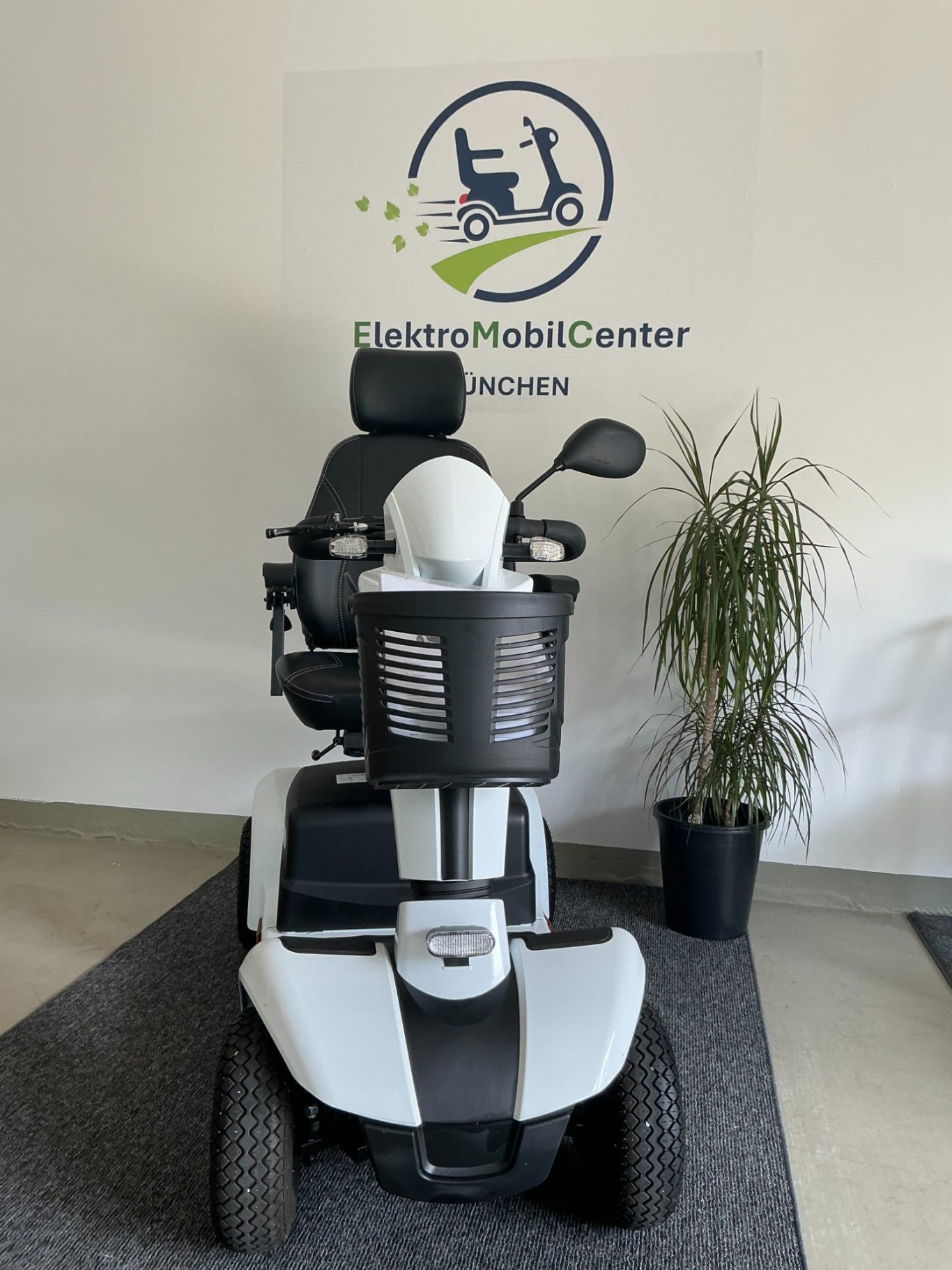 Rollstuhl mit Elektroantrieb, vor einem Unternehmenslogo und einer Pflanze platziert.