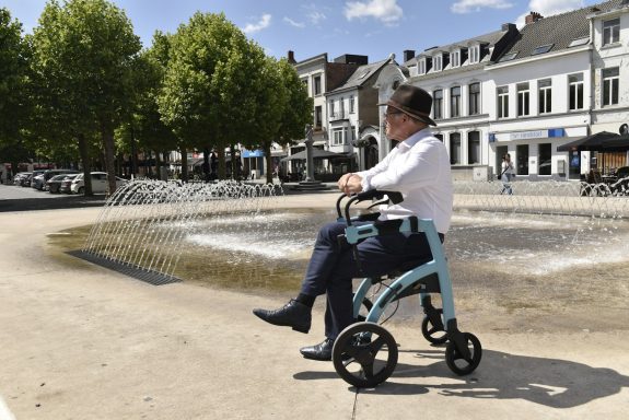 Person mit Rollator fährt an einem Brunnen auf einem Platz vorbei, umgeben von Bäumen.