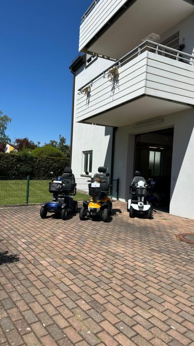 Drei Mobilitätsroller stehen vor einem modernen Gebäude auf gepflastertem Boden.